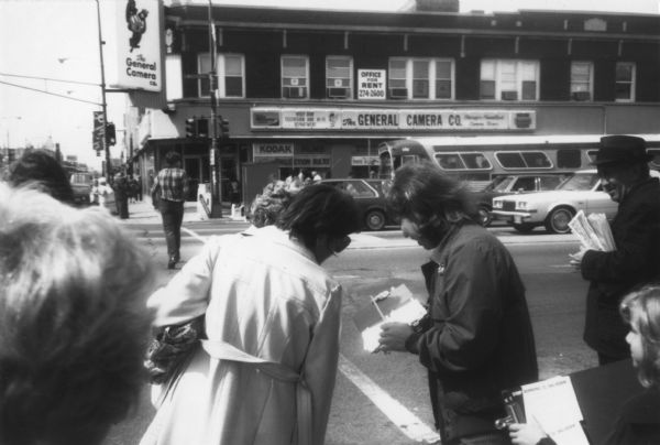 A woman is standing on a street corner showing a piece of paper to two people. Across the street are signs for The General Camera Co. Caption reads: "Gayle McLaughlin getting cards signed." A woman in the foreground on the right is holding a clipboard with cards that read: "Stop Bombing El Salvador."