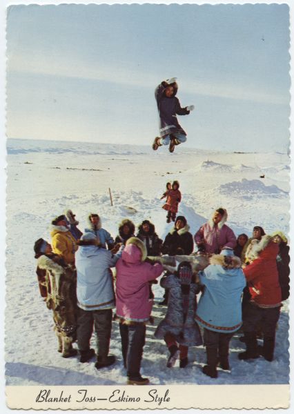 Slightly elevated view of a group of people bundled up in colorful coats, standing in a snowy landscape. They are all holding one blanket and looking up towards a smiling individual in the air. Behind the big group are two children standing on a pile of snow. Text at bottom reads: "Blanket Toss — Eskimo Style." On the reverse side of postcard text at top reads: "Another -- 'alaska joe' original." At bottom left text reads: "Originally hunters were elevated this way to see over ice ridges to spot game. Alaska Airlines Photograph." Handwritten message reads: "Anchorage, Alaska. July 21 Dear Eleanor - How are you this summer. Hope you are having good weather. Today is beautiful here. Wish you could see the many flowers. We saw a blanket toss when we were in Kotzebue. With best wishes to you and Davis. Josephine."