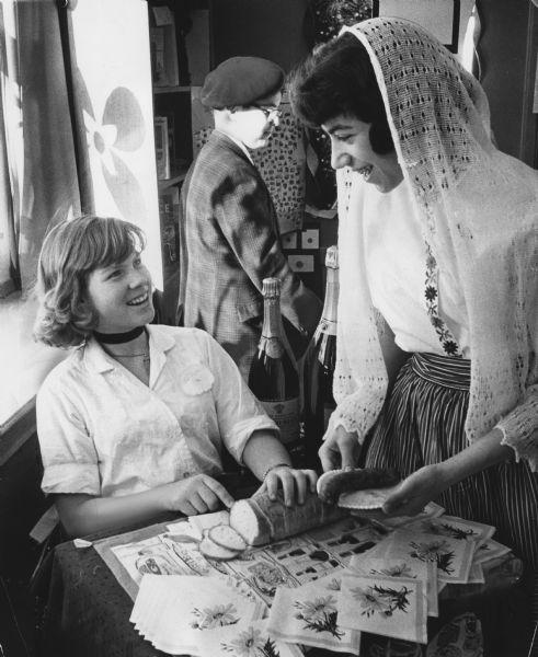 Three students at folk fair. One student is sitting at a table slicing bread, with a student standing next to her who is holding a sausage on a plate. A third student wearing a beret is standing behind them. There are two bottles of champagne and flower printed napkins on the table. Text reads: "Pupils at Cumberland school in Whitefish Bay, Cheryl Olsen (left), 4890 N. Lake dr., and Nancy Kaufer, 13, of 4839 N. Elkhart av., sampled refreshments Tuesday at a continental folk fair held at the school. The junior high program featured decorations, costumes and food representing various European countries. —Journal Photo"