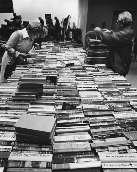 View down table that is packed with books towards people standing on either side examining the titles. Caption reads: "READER'S DELIGHT — Prospective buyers browsed through the used books offered at Northridge Shopping Center Wednesday by the Milwaukee book sale committee of the Brandeis University Library National Women's Committee. Proceeds from the sale, which continues through Friday in the center's Community Room, will be used to buy books for the university, which is in Boston, Mass."