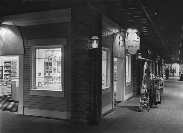 A woman is pushing a stroller with an infant in it while holding clothes wrapped in plastic. She is walking down a brick paved corridor near a storefront designed to look like the exterior of an older building, with a sign hanging above the doorway, and a lamppost at the corner. Caption reads: "The County Faire Shopping Center in Grafton, with its look of the 1890s, is located on a 24 acre site."