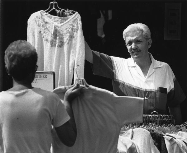 A man is holding up a sweater on a hangar over a clothing rack. in the foreground a woman is facing him and holding up another shirt. Caption reads: "Terry Soykup of Brookfield compared shirts while shopping with his wife at the sidewalk sale."