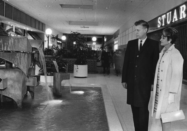 A man and woman are standing and looking at a water fountain with an abstract art sculpture in it. Caption reads: "<b>Visions of decorations</b> dance in the heads of Mr. and Mrs. Edward A. Grede as they cast appraising eyes at the fountain in Brookfield Square, where the first Opera club ball will be held Sept. 21. Mr. and Mrs. Grede, 1650 Shady la., Elm Grove, are chairmen of the benefit for the Florentine Opera Co."