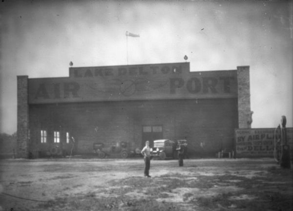 Two men are standing in front of the Lake Delton airport hangar with a windsock on the roof. Automobiles are parked inside the hangar. On the right is a fuel pump, and behind it near the hangar is a large sign that reads: "Scenic Excursion [obscured] By Airplane," and "Tips Over Dells." 