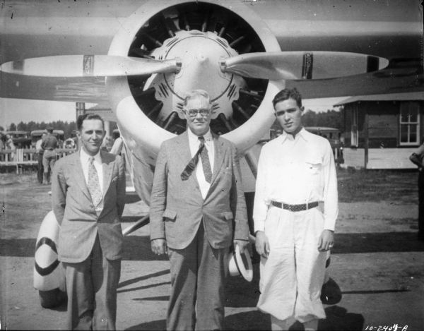 Group portrait of three men: the Lake Delton airport owner, likely William J. Newman (center), manager and son standing in front of the prop of an airplane. People are standing in the background in front of a fence, and behind the fence are parked automobiles.
