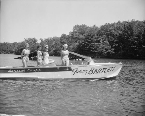 Four women are standing in the Tommy Bartlett boat. Three of them are wearing striped bathing suits, and one woman is wearing a white bathing suit. Two water skiing jumping ramps are behind the boat.