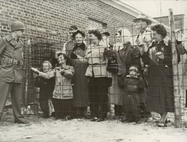 Caption reads: "Citizens of Mosinee who could not show proper credentials were thrown into the stockade. Photo shows women pleading for food as armed guard stands outside fence. INP Photo by F. Stanfield." Caption on back is a newspaper clipping that reads: "Men, women and children of Mosinee know now what happens when you don't have the proper credentials in Communist-controlled countries. This group landed behind a barbed-wire fence, and pleading with the trooper (left) was like talking to a wall. He had a heart of stone. Sentinel photo."