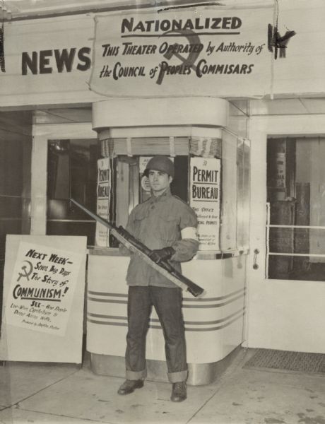 Caption reads: "Simulated Communist Control-The local movie theater is seized by Communist authorities and pictures confiscated with Communist dominated films substituted. Armed guard stands at entrance." Caption on back is a newspaper clipping that reads: "Moving picture films at the Mosinee theater were confiscated and the Communists replaced them with their own propaganda films. Sentinel photo."