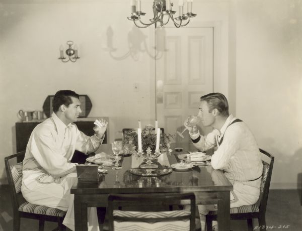 Cary Grant and Randolph Scott sitting opposite each other at a dining table in their house. Grant is holding a sandwich in one hand, while Scott has a glass raised to his mouth as they look directly at one another.