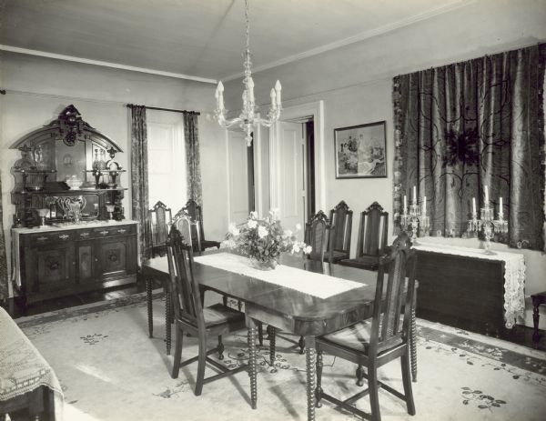 Interior view of the dining room from the kitchen doorway. There are two doors on the opposite side of the room: the one on the left leads to the parlor, and the one on the right leads to the stairwell. There is a large sideboard on the wall on the left, and a painting and a wall hanging are on the right above a table with two large glass candelabras.