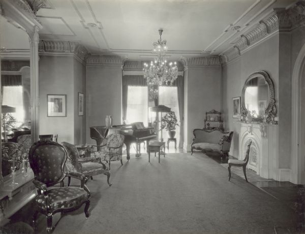 Interior view of the parlor in the Governor's Mansion. A large mirror is on the wall on the left, chairs are arranged along the left side of the room, and a piano is near the large windows in the background, which faces the street. There appears to be a tall, narrow mirror between the windows, reflecting the chandelier in the center of the room. On the right near the arched doorway, which leads to the hall, is a fireplace with a large mirror on the mantle.