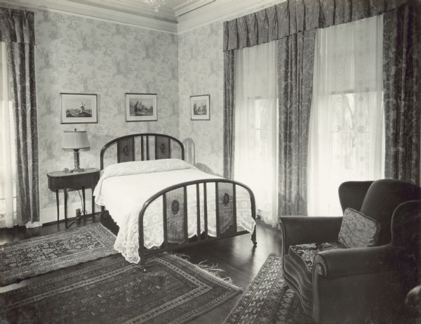 A bedroom on the second floor of the mansion. The window on the left  of the bed faces the street, and there are two windows on the right wall. Three rugs are on the floor, and three framed prints are hanging on the wall above the bed in the corner. An upholstered chair is in the foreground on the right.