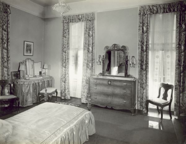 The front bedroom on the second floor of the mansion. The foot of the bed is in the foreground, and in the corner on the left near a window is a dressing table with a mirror and a bench. A dresser with a mirror is on the far wall between two windows that face the left side yard of the property. The mirror is reflecting the headboard of the bed, a door frame, and the shower curtain in the bathroom.