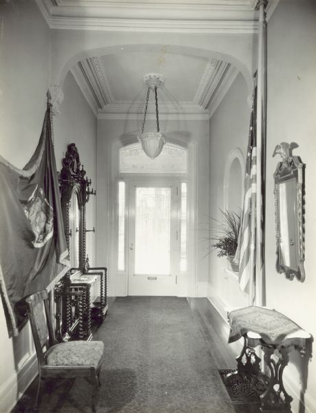 View of the entrance hall looking towards the front door, which has sidelights, and a fan window above covered by a curtain. On the left is a large carved wood hall tree with a mirror. In the foreground on the left is a Wisconsin flag, and on the right a United States flag. A plant is in an arched niche behind the flag on the right.