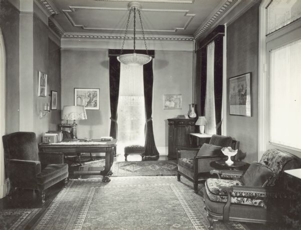 Drawing room on the first floor in the Governor's Mansion. On the left is a chair near the arched doorway that leads to the hall. There is a desk in the left corner of the room. A window with dark-colored drapes and a lace curtain is along the back wall which faces the street. On the right along the wall is a chair near a window with matching dark-colored drapes. In the right foreground is a chair near a door to the porch which has curtains over the window and fan window.