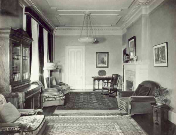 Drawing room in the Governor's Mansion. On the left is a chair near a bookcase, and two more chairs in front of windows with dark-colored drapes and lace curtains. There is a closed door on the back wall on the left which leads to the dining room. A curtained doorway on the right wall in the corner leads to a short hall under the stairs. Along the right wall are two chairs near a fireplace.