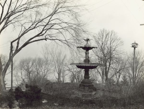 View of the fountain, which is from the second Capital, in the backyard. Lake Mendota is down the hill in the background. The fountain is empty, and the trees are bare of leaves. There are birdhouses on posts on the left and right.