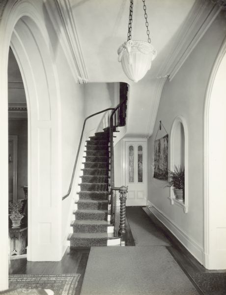Front entry looking towards curved staircase. An arched doorway leading to the Drawing Room is on the left, and the arched doorway on the right leads to the Parlor. Along the wall on the right is a potted plant in an arched alcove and a tapestry wall hanging. A closed door with two leaded glass windows is in the back under the staircase, which leads to the Dining Room.