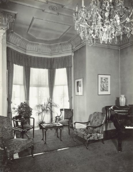 Interior view of the alcove, with three bay windows which have tied back drapes, a valance, and lace curtains. The view looks out to the right side yard of the property. Four chairs, a small table, and two potted plants are in the alcove. On the right is a grand piano in the corner of the room, and a chandelier is in the center foreground.