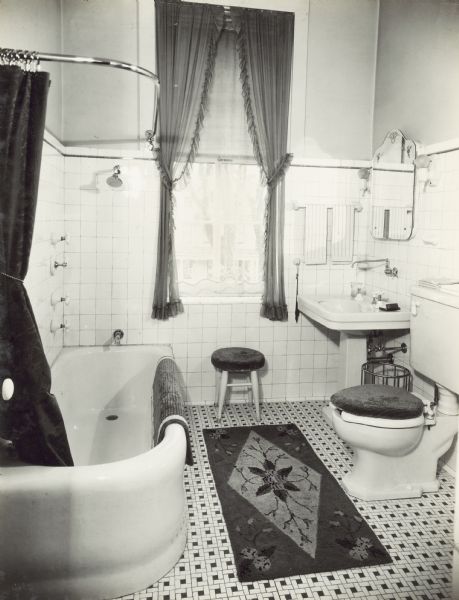 Bathroom on second floor. The window looks out onto Gilman Street. There is a rug on the tile floor, and the shower curtain has been pulled back by a tie. The plumbing fixtures in the room, which include a tub, shower head, sink and toilet, are Kohler.
