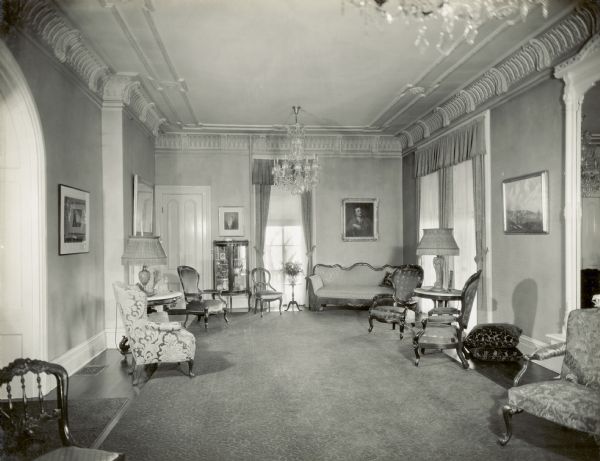 A longer view of rear of the Parlor, with a view of the arched doorway on the left that leads to the front hall, and a large mirror on the wall on the right. The door in the left corner is closed. The window on the back wall looks out towards Lake Mendota, and has the original 1850 window sash.