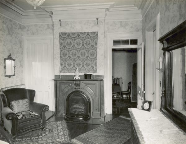 Master bedroom with fireplace flanked by two doors. There is a chair in the left corner near a closed door. On the right is an open door with a transom leading to a hallway where there is a chair next to a telephone on a table. Along the right in the foreground is a dresser with a mirror, and a closed closet door. There is a wall hanging above the mantle, and rugs on the floor.