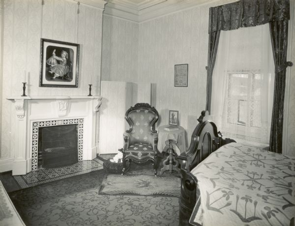 The bed is in the right foreground and is covered with a quilt. A chair, table, and floor screen are in the corner in the center, with a window on the right with a view of Lake Mendota. There is a fireplace along the left wall. A framed portrait of Ole Bull is on the table. 