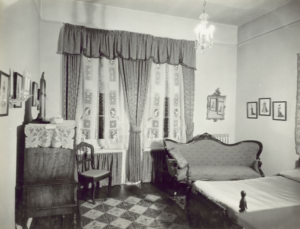 Bedroom with a bed on the right wall, with a wall hanging above it. There is a chair and settee in front of a double window along the back wall, which looks out towards the left side yard of the property. The windows have tie-back drapes and a valance, and matching drapes below the window sills. There is a dresser with a small mirror along the wall on the left, and framed images and a mirror are along the walls.
