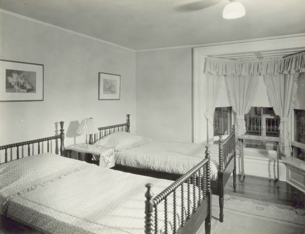 Bedroom on second floor, with a bay window looking out into the side yard, and leading to the balcony. There are two beds, with a table and lamp between them, along the wall on the left. 