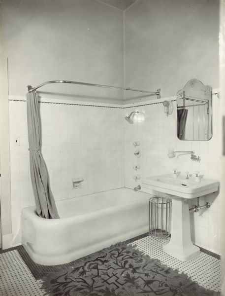 Bathroom (Jack and Jill style), with a view of the bathtub, shower head, and sink. The shower curtain has been pulled back. There is a rug on the tile floor, and a door frame is to the left of the tub.