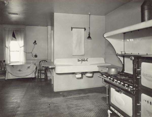 View of the kitchen with a Reliant stove in the foreground along the right wall. A large sink is along a wall just beyond the stove. In the background on the left is a table and two chairs in front of a window with an exhaust fan at the top. The window looks out into the right side yard of the property. A door frame in the center leads to a room with a cupboard behind the sink.