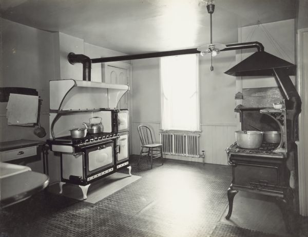 Another view of the kitchen, with the Reliable stove against the wall on the left, and a corner of the sink in the foreground. The window on the back wall has a view of the left side yard. The door in the corner behind the stove leads to the dining room. There is another stove against the right wall corner near a door frame, which leads to the back stairway.