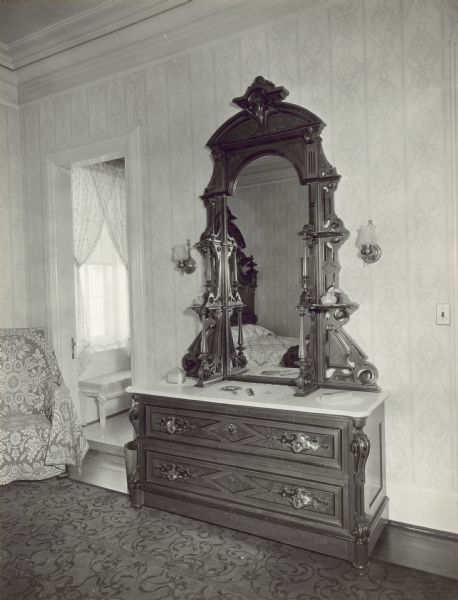 A dresser with a large, wood-framed mirror is along the wall, and a bed covered with a quilt is reflected in the mirror. In the left corner of the room is a chair next to an open door leading to the Jack and Jill style bathroom. The window in the bathroom looks out to the right side yard of the property. 