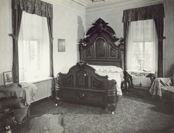 Right rear master bedroom with a large, carved wood bed with tall headboard set at an angle in the corner of the room. There is a framed portrait of Ole Bull on the table on the left, which is next to a window above a radiator. The window looks out towards Lake Mendota. The window on the back right wall looks out onto the right side yard.