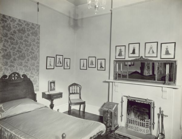 On the left is a bed with a headboard against the wall, with a wall hanging above it. There is a chair in the corner in the center, and on the right wall is a fireplace. Framed images are along the wall and above the mirror on the mantle. Reflected in the mirror is a settee, and  two windows with drapes and lace curtains.
