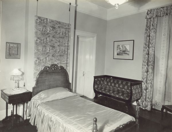 A small table is to the left of the bed, which has a headboard, and a wall hanging above it. There is an inset closed door to the right which leads to the front bathroom. Along the wall near the door is a settee, with a window on the right which looks out towards Gilman Street.