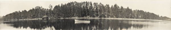 Panoramic view across water towards the Trout Lake shoreline. There are two men in a boat in the water in the foreground. There is a boathouse on the shore on the left. In the center among trees is the Main Building.