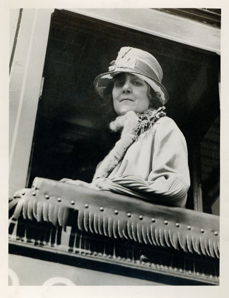 Zona Gale is looking down from a balcony lined with fringe. She is wearing a hat and coat.