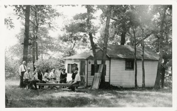 Members of the Western Wisconsin Camp Association are gathered outside of a cabin sitting on benches.  