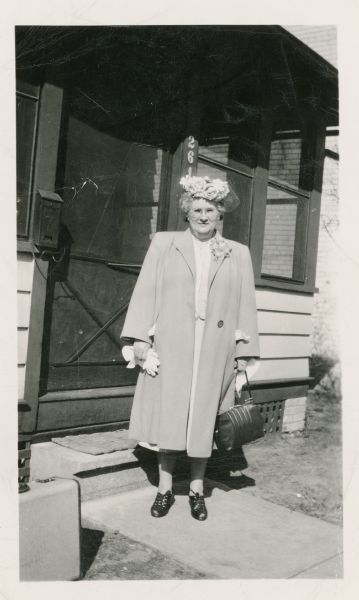 Melvina Hostak posing outside of a house in front of a door. She is wearing a coat and hat, and is holding gloves in one hand, and a handbag in the other hand.