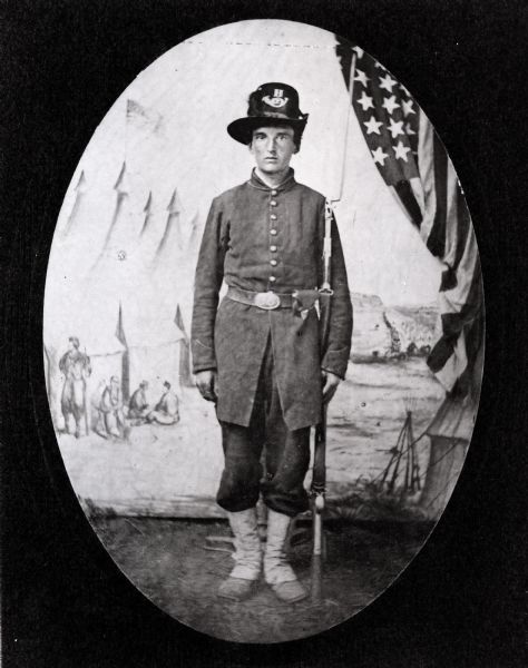 Full-length portrait in front of a painted backdrop and a U.S. Flag of Civil War soldier Elon Frances Brown of the 2nd Wisconsin Infantry, posing in uniform. He is holding a rifle with a bayonet.