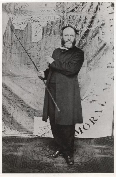 Portrait of Reverend Edward Mathews standing in front of a "Moral Map of the United States." He is holding a long, wooden pointer and is gesturing towards the map.