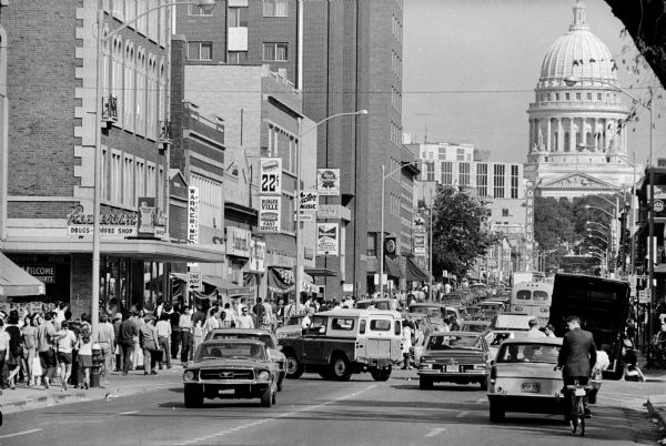 A view up State Street from the corner of Lake and State Streets showing heavy automobile and pedestrian traffic. The Wisconsin State Capitol and the Orpheum Theater are in the background. A number of State Street businesses are on the north side of the street, including Rennebohm's, Burger Ville, Victor Music, The Grotto restaurant, and The Pub.