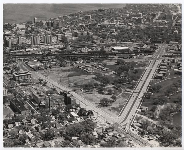 Air view looking north toward Lake Mendota. The intersection of West Washington Ave. and S. Park Street is at bottom center. The Triangle Redevelopment Project is underway. No buildings from the Greenbush neighborhood remain and pedestrian overpasses cross both streets. Meriter Hospital and Lowell School are at bottom left. A rail line crosses near center with a portion of the roundhouse remaining right of center. The Wisconsin State Capitol is at top right and several University dorms are visible near top left.