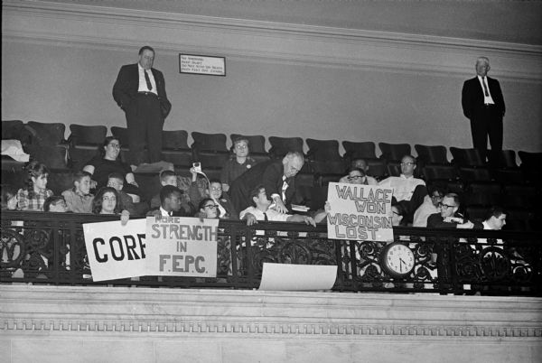 Congress on Racial Equality (CORE) protestors with signs sitting in the balcony of the Wisconsin Assembly Chambers. Assembly sergeant-at-arms, Norris Kellerman is reaching to remove a sign on the order of the speaker at the request of a Democratic Assembly member from Milwaukee. Two other men in suits are standing in the background. No signs are allowed in the Wisconsin Assembly Chambers.