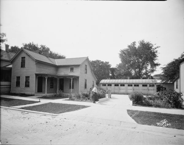 View from street towards a house and flower garden at 614 E. Main Street. There is a man (possibly Emanuel F. Yager) and woman standing in the driveway with a Trachte 4 bay garage in the background. 