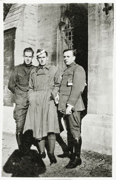 Three men in military uniforms are posing together in front of a stone building. 