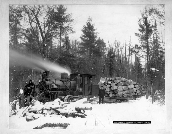 Several man posing by a steam skidder which is hauling logs.