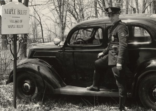A uniformed officer standing and posing with one leg on the running board of an automobile parked near a sign that reads: "Maple Bluff." He is wearing a Traffic Police badge and a service revolver. He is identified in a caption as Marshall [A.J.] Taff of Maple Bluff.