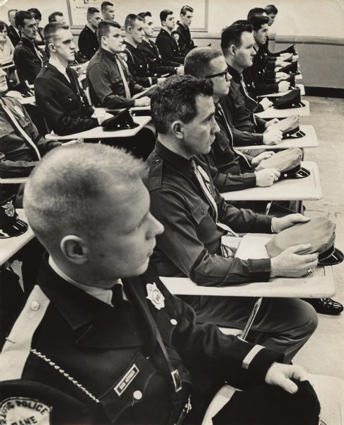 View of a group of uniformed officers are sitting in rows at desks with their hats set on the desktop in front of them. The closest officer has a Dane Traffic Police patch on his sleeve.